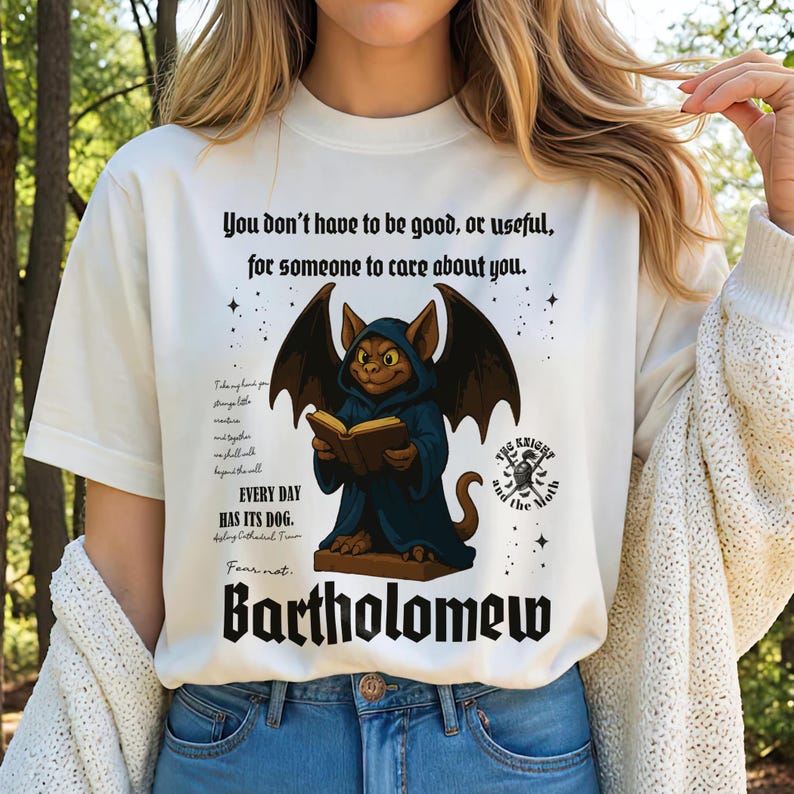 May include: Cream-colored t-shirt featuring a cartoon illustration of a creature with bat wings, reading a book. The text on the shirt reads "You don't have to be good, or useful, for someone to care about you." and "Bartholomew."