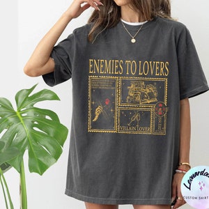 Puede incluir: Una camiseta gris oscuro con el texto "Enemies to Lovers" impreso en dorado. El dise&ntilde;o presenta una serie de sellos de estilo vintage con ilustraciones y texto, incluyendo "Villain Lover" y "Issued".