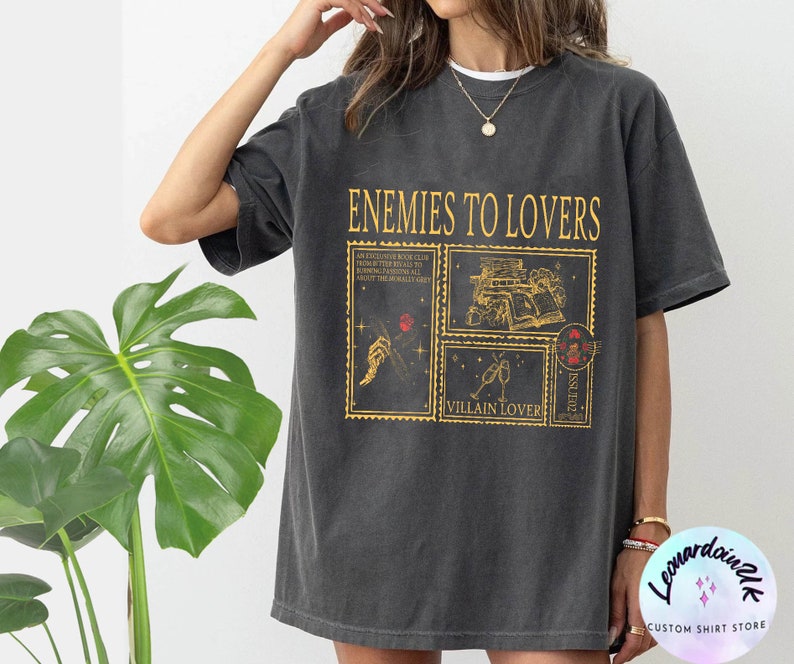 Puede incluir: Una camiseta gris oscuro con el texto "Enemies to Lovers" impreso en dorado. El dise&ntilde;o presenta una serie de sellos de estilo vintage con ilustraciones y texto, incluyendo "Villain Lover" y "Issued".