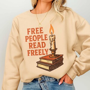 May include: Beige sweatshirt featuring the orange text "FREE PEOPLE READ FREELY". A candle with a lit flame is positioned above two books. The design has a vintage, retro aesthetic.