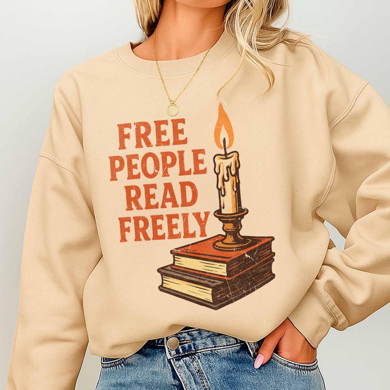 May include: Beige sweatshirt featuring the orange text "FREE PEOPLE READ FREELY". A candle with a lit flame is positioned above two books. The design has a vintage, retro aesthetic.
