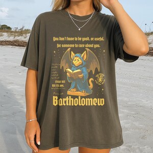May include: Dark olive green t-shirt featuring a cartoon illustration of a blue creature with bat wings reading a book. The text on the shirt reads "You don't have to be good, or useful, for someone to care about you." and "Bartholomew."