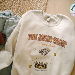 May include: A cream colored sweatshirt with a graphic print of a hand reaching down to a crown with the text "The Russo Group Luxury Goods" and "Dante Russo" below the crown.
