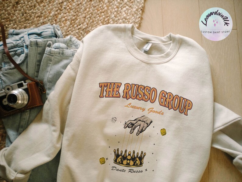 May include: A cream colored sweatshirt with a graphic print of a hand reaching down to a crown with the text "The Russo Group Luxury Goods" and "Dante Russo" below the crown.