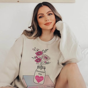 May include: A cream-colored sweatshirt with a graphic of a mason jar filled with pink and white flowers. The jar is sitting on a book with the text "a thousand boy kisses" and "Tillie Cole" printed on the cover.