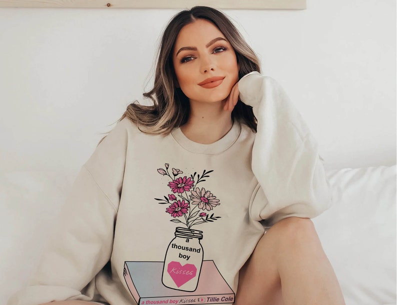 May include: A cream-colored sweatshirt with a graphic of a mason jar filled with pink and white flowers. The jar is sitting on a book with the text "a thousand boy kisses" and "Tillie Cole" printed on the cover.