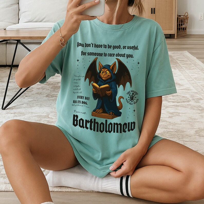 May include: A light teal t-shirt featuring a cartoon illustration of a creature reading a book, with the name "Bartholomew" below. The shirt also has the text "You don't have to be good, or useful, for someone to care about you."