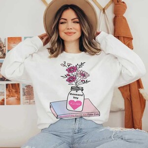 May include: White sweatshirt with a graphic of a mason jar filled with pink and purple flowers. The jar is sitting on top of a book with the title "A Thousand Boy Kisses" by Tillie Cole.