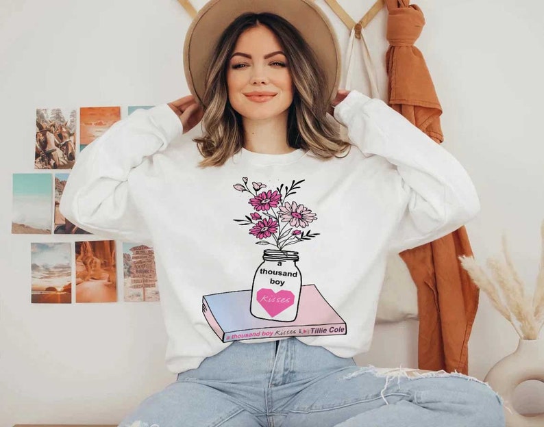 May include: White sweatshirt with a graphic of a mason jar filled with pink and purple flowers. The jar is sitting on top of a book with the title "A Thousand Boy Kisses" by Tillie Cole.