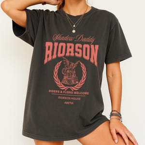 May include: Dark grey t-shirt with a distressed appearance. The front displays the text "Shadow Daddy RIORSON" in a red, vintage font, accompanied by a dragon and castle graphic. Below, it reads "Riders & Fliers Welcome, Riorosn House, Aretia."