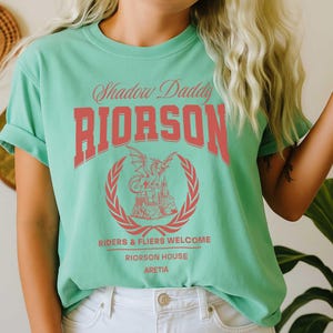 May include: A pale green t-shirt featuring the red text "Shadow Daddy RIORSON" and a dragon and castle emblem. The shirt also displays the phrases "RIDERS & FLIERS WELCOME" and "RIORSON HOUSE ARETIA".