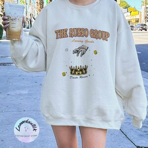 May include: A white sweatshirt with a graphic design featuring the text "THE RUSSO GROUP" in orange and brown lettering. The graphic also includes a hand holding strings attached to a gold crown with the text "Luxury Goods" and "Dante Russo" below the crown.