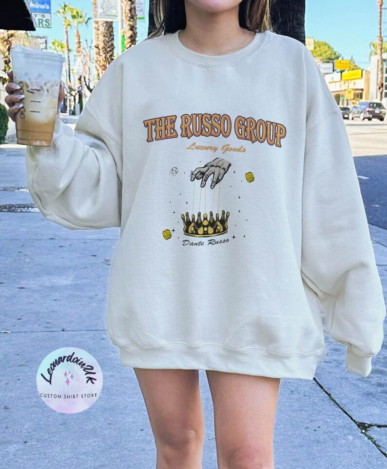 May include: A white sweatshirt with a graphic design featuring the text "THE RUSSO GROUP" in orange and brown lettering. The graphic also includes a hand holding strings attached to a gold crown with the text "Luxury Goods" and "Dante Russo" below the crown.
