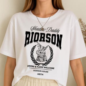 May include: White t-shirt featuring a black graphic. The design includes the text "Shadow Daddy RIORSON" above a crest with a dragon and castle illustration. Below the crest, it reads "RIDERS & FLIERS WELCOME RIORSON HOUSE ARETIA".