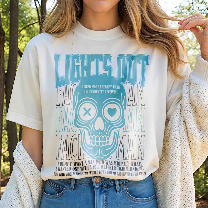 Pu&ograve; includere: T-shirt color crema con grafica di teschio blu e la scritta "LIGHTS OUT". La grafica include il testo "I NEED MORE THERAPY THAN I'M CURRENTLY RECEIVING" e una citazione sul desiderio di un'anima pi&ugrave; oscura.