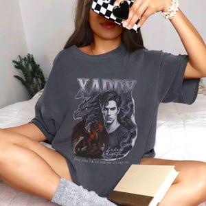 Puede incluir: Camiseta gris oscuro con un dise&ntilde;o gr&aacute;fico. El dise&ntilde;o incluye la palabra "XADDY" sobre una ilustraci&oacute;n de un hombre y dos dragones. El texto "Even when I'm not with you, it's only you" est&aacute; impreso debajo. La camiseta tiene un corte relajado.