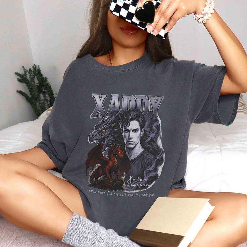 Puede incluir: Camiseta gris oscuro con un dise&ntilde;o gr&aacute;fico. El dise&ntilde;o incluye la palabra "XADDY" sobre una ilustraci&oacute;n de un hombre y dos dragones. El texto "Even when I'm not with you, it's only you" est&aacute; impreso debajo. La camiseta tiene un corte relajado.