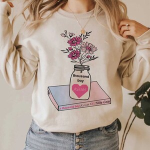 May include: A cream-colored sweatshirt with a graphic of a mason jar filled with pink flowers. The jar is labeled "a thousand boy kisses" and sits on top of a book with the title "a thousand boy kisses by Tillie Cole".
