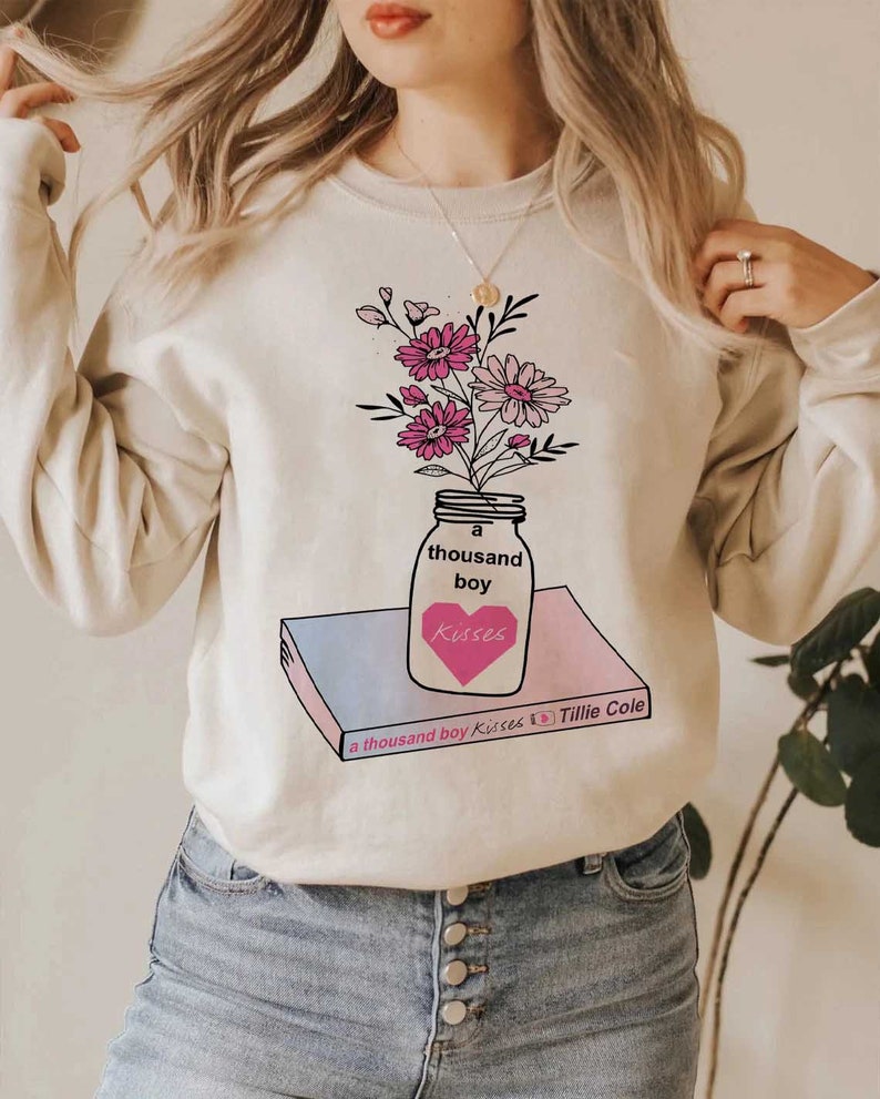 May include: A cream-colored sweatshirt with a graphic of a mason jar filled with pink flowers. The jar is labeled "a thousand boy kisses" and sits on top of a book with the title "a thousand boy kisses by Tillie Cole".