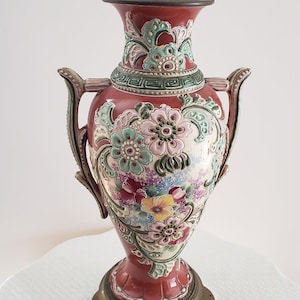 May include: A ceramic table lamp featuring a red body with raised floral relief patterns in shades of pink, green, and yellow. The lamp has two side handles, a Greek key border design, and sits on a circular, ornate, dark metallic base.