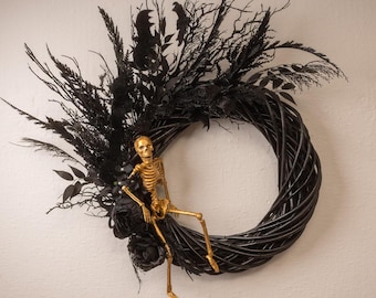 May include: A black wicker wreath decorated with black feathers, leaves, and a gold skeleton sitting on the wreath.