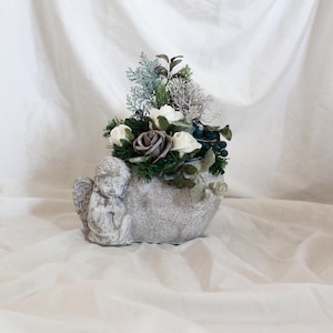 May include: A gray, stone-like planter shaped like a bowl with an angel figurine on the side. The planter is filled with white and gray roses, greenery, and small blue berries. The background is a white fabric.