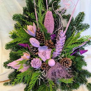 Puede incluir: Arreglo floral decorativo con ramas de hoja perenne, piñas y diversas flores secas en tonos verdes, morados y rosas. El arreglo presenta una variedad de texturas y formas, creando una exhibición visualmente atractiva.