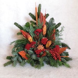 Könnte beinhalten: Dekorative Blumengesteck mit grünen Tannenzweigen, roten Beeren und getrockneten orangefarbenen Blättern und Samenkapseln. Die Anordnung ist dreieckig und eignet sich für einen Tischschmuck oder eine saisonale Dekoration.