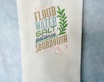 Op de afbeelding: Witte keukendoek met geborduurde tekst: "FLOUR WATER SALT patience SOURDOUGH" in bruin, blauw en groen. Een groene blad tak is naast de tekst geborduurd. De doek is gemaakt van een lichtgekleurde stof.