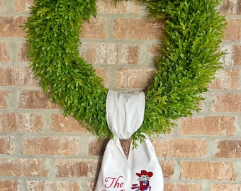 May include: A green, leafy wreath with a white ribbon featuring the embroidered text "The Belks" and a cartoon image of a person in a cowboy hat. The wreath hangs against a brick wall.