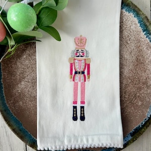 May include: White hand towel with an embroidered pink and gold nutcracker soldier design. The towel has a pom-pom trim and is displayed on a decorative plate.