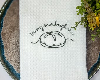 Op de afbeelding: Witte keukenhanddoek met wafelweefsel met de geborduurde zin "in my sourdough era" en een broodontwerp. De handdoek ligt op een decoratieve schaal met een blauwe en bruine rand, met groen en een gouden vlinderaccent.