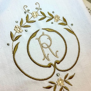 May include: A white embroidered kitchen towel with a gold monogram of the letter "R" surrounded by a wreath of green leaves and white flowers.