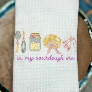 May include: White waffle-weave kitchen towel embroidered with a spoon, whisk, jar, loaf of bread, and a bundle of flowers. The phrase "in my sourdough era" is embroidered in pink script. The towel is folded and placed on a blue and brown plate.