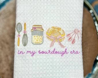 Op de afbeelding: Witte keukenhanddoek met wafelweefsel, geborduurd met een lepel, garde, pot, brood en een bos bloemen. De zin "in my sourdough era" is geborduurd in roze script. De handdoek is opgevouwen en op een blauw-bruin bord geplaatst.