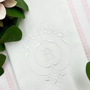 Può includere: Un tovagliolo di lino bianco con un ricamo floreale e la lettera "R" al centro. Il ricamo è in filo bianco, creando un aspetto delicato ed elegante. Il tovagliolo è piegato e posto su una superficie a righe rosa e bianche.