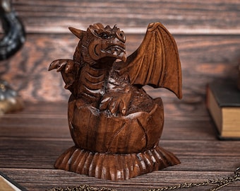 May include: A hand-carved wooden figurine depicting a small dragon emerging from a cracked egg. The sculpture features detailed scales, wings, and facial features in a rich, dark brown wood grain finish, standing on a textured, flared base.