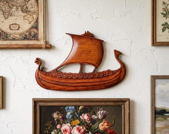 May include: A carved wooden wall hanging shaped like a Viking longship. The piece features a prominent curved sail, a dragon-headed prow, and a series of circular shield motifs along the side, all finished in a warm, polished brown wood grain.
