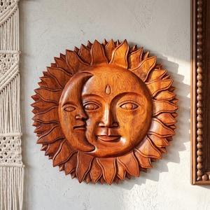 Puede incluir: Una talla de pared circular de madera que presenta un rostro de sol con una luna creciente superpuesta en el lado izquierdo. La pieza tiene un acabado marrón cálido con vetas de madera visibles y rayos puntiagudos hacia el exterior.