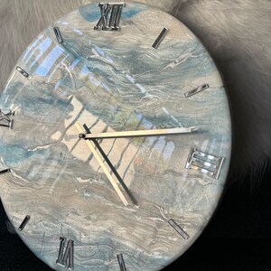 Puede incluir: Un reloj redondo con un dise&ntilde;o de m&aacute;rmol azul y gris. La esfera del reloj tiene marcas de n&uacute;meros romanos y manecillas plateadas.