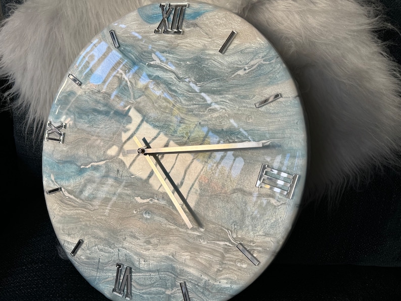 Puede incluir: Un reloj redondo con un dise&ntilde;o de m&aacute;rmol azul y gris. La esfera del reloj tiene marcas de n&uacute;meros romanos y manecillas plateadas.