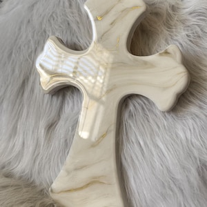 May include: A decorative cross with a marble-like appearance. The cross is primarily white with gold veining and accents. The cross has a smooth, glossy finish and is set against a soft, gray background.