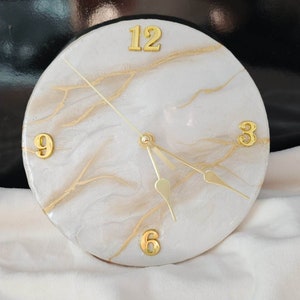 May include: White and gold resin clock with Roman numeral markings. The clock face has a marbled design with gold accents.