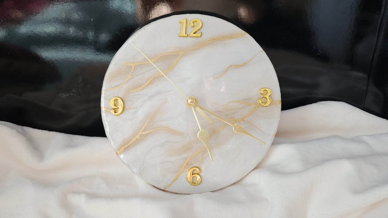 May include: White and gold resin clock with Roman numeral markings. The clock face has a marbled design with gold accents.