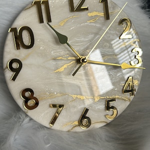 May include: A white and gold resin clock with gold Roman numeral hour markers and gold hands. The clock face has a marbled design with gold accents.