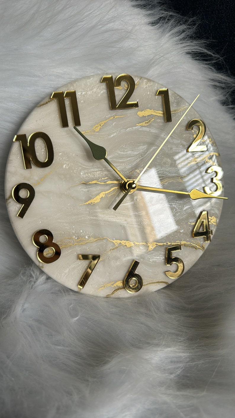 May include: A white and gold resin clock with gold Roman numeral hour markers and gold hands. The clock face has a marbled design with gold accents.