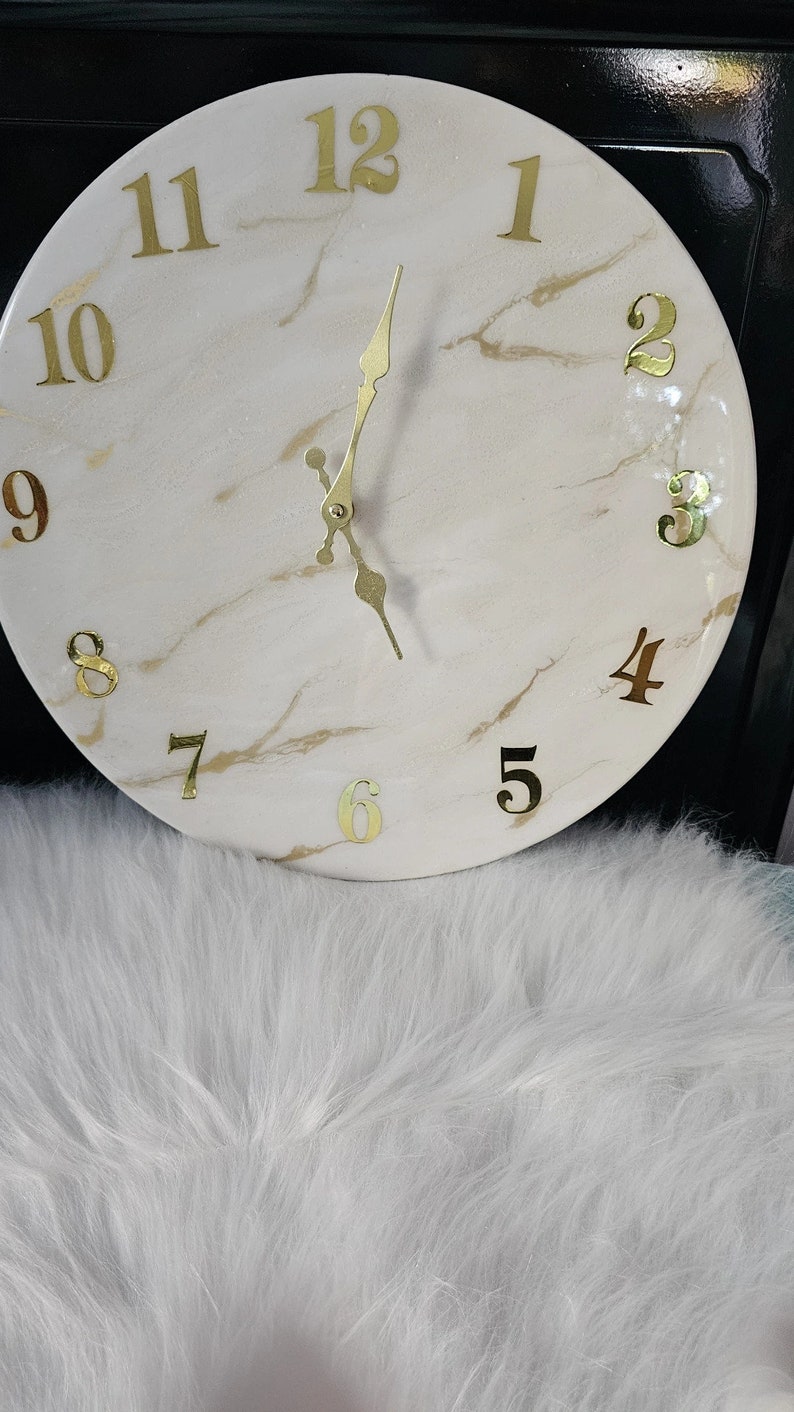 May include: A white clock with gold numbers and hands. The clock face has a marble-like design.
