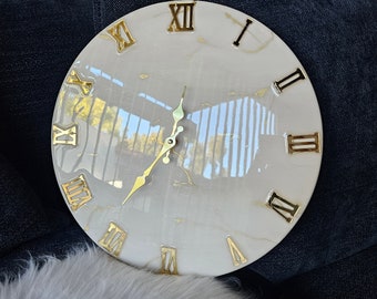 May include: A white round clock with gold Roman numeral markings and gold hands. The clock face has a marble-like pattern with gold flecks.