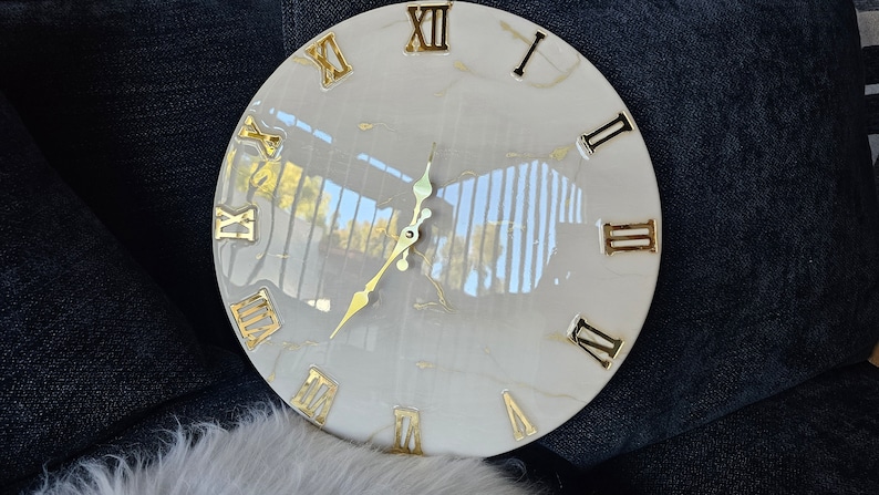 May include: A white round clock with gold Roman numeral markings and gold hands. The clock face has a marble-like pattern with gold flecks.