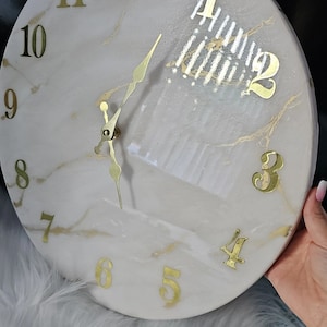 May include: A white and gold marble clock with gold Roman numeral hour markers and gold hands. The clock face has a white background with gold marble accents.